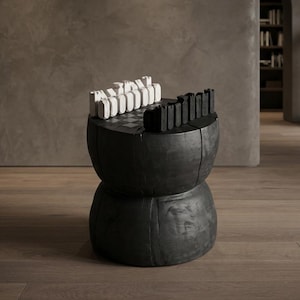 Può includere: Un tavolo a piedistallo in legno nero carbone con una base cilindrica a forma di clessidra. La superficie superiore presenta una scacchiera integrata con un set di pezzi bianchi e neri disposti in file sui lati opposti del tabellone.