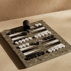 May include: A backgammon set with a speckled grey board, black and white playing pieces, and a black dice cup. The board features black and white triangles and is set on a light-coloured surface.