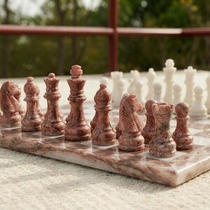 Puede incluir: Un juego de ajedrez con piezas de piedra pulida en tonos rosa y blanco. El tablero est&aacute; construido con cuadrados de m&aacute;rmol rosa y blanco a juego. El conjunto incluye piezas talladas de rey, reina, alfil, caballo, torre y pe&oacute;n sobre el tablero.