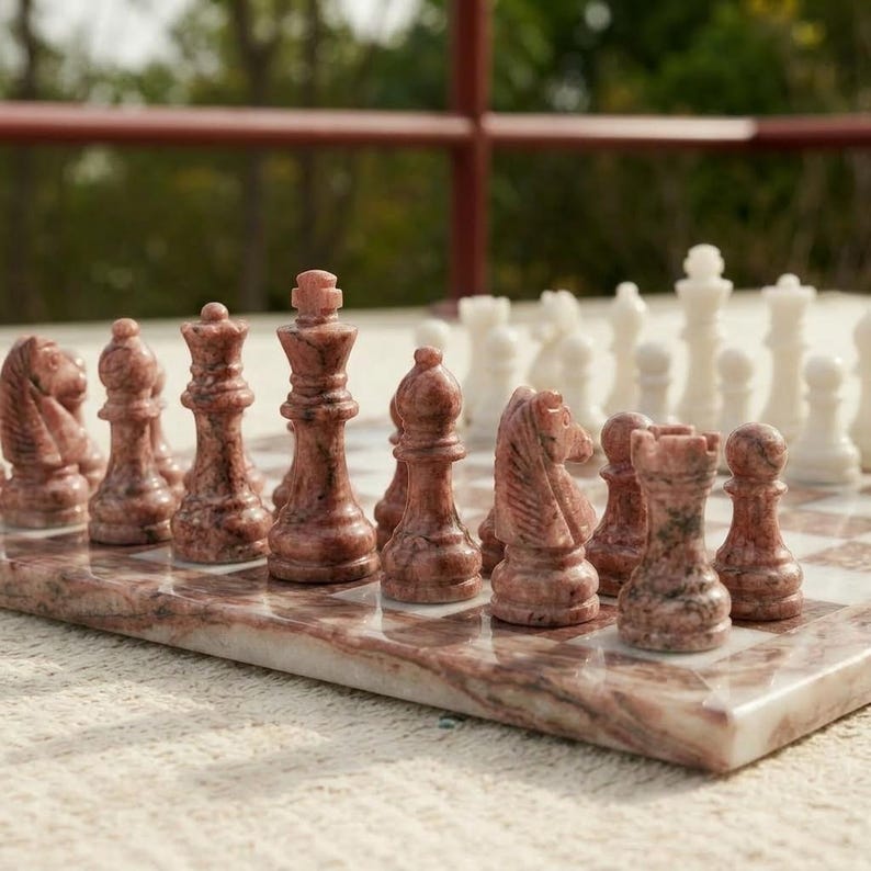 Puede incluir: Un juego de ajedrez con piezas de piedra pulida en tonos rosa y blanco. El tablero est&aacute; construido con cuadrados de m&aacute;rmol rosa y blanco a juego. El conjunto incluye piezas talladas de rey, reina, alfil, caballo, torre y pe&oacute;n sobre el tablero.