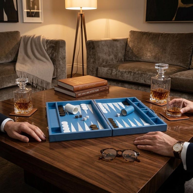 May include: A blue backgammon set with white and brown game pieces is open on a dark wood table. Two crystal decanters and a glass of amber liquid sit on the table. A pair of eyeglasses rests on the table.