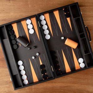 Può includere: Un set da backgammon nero con dettagli arancioni e bianchi. La tavola da gioco aperta mostra triangoli arancioni, pedine bianche e nere, dadi e bicchieri per dadi. Il set è disposto su una superficie di legno.