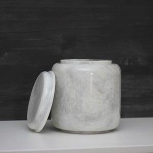 May include: A white marble jar with a matching lid. The jar is cylindrical with rounded edges and a wider base. The lid is flat and circular. The marble has a subtle, swirling pattern. The jar and lid are on a white surface.
