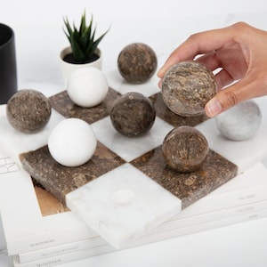 May include: A marble checkerboard game set with brown and white marble pieces. The board is made of white and brown marble squares. The pieces are arranged on the board in a game in progress.
