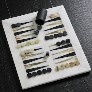 May include: A white marble backgammon board with black and white playing pieces. The board has a pattern of black and white triangles and green and brown squares. There are two black dice on the board, one showing a "4" and the other showing a "6".