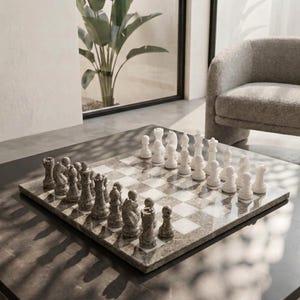 May include: A square chess board featuring a checkered pattern of polished gray granite and white marble squares. The board is set with a complete collection of carved chess pieces, with one side in speckled gray stone and the other in solid white stone.