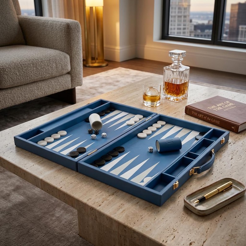 May include: A blue backgammon set with white and black game pieces, dice, and a dice cup. The set is open on a stone table, with a decanter, a glass of amber liquid, and a book titled "The Art of the Game" nearby. A pen and tray are also on the table.