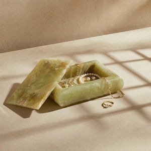 Puede incluir: Caja de joyería rectangular de ónix verde abierta con tapa con bisagras. En el interior, se ven collares de oro, una pulsera de perlas y un anillo. La caja está sobre una superficie clara.