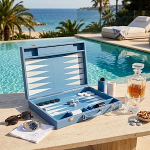 Puede incluir: Un juego de backgammon azul claro con piezas blancas y negras, dados y un asa. Un reloj, gafas de sol y un vaso de licor están cerca. El juego está abierto, revelando el tablero.