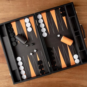 May include: A black backgammon set with orange and white accents. The game board is open, revealing a playing surface with orange triangles and white and black game pieces. Dice and dice cups are also visible, set on a wooden surface.