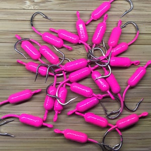 May include: A pile of pink fishing jig heads with silver hooks. The jig heads are shaped like a cylinder and have a small eyelet at the top for attaching a fishing line.