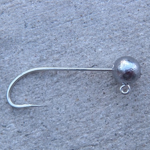 May include: A silver-colored fishing jig head with a round weight and a curved hook. The hook has a sharp point and a small eyelet for attaching a fishing line. The jig head is designed for use in freshwater or saltwater fishing.