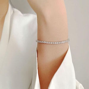 May include: A tennis bracelet featuring a row of small, round, clear gemstones. The bracelet is silver-toned and worn on a wrist, against a white shirt. The gemstones are evenly spaced and reflect light.