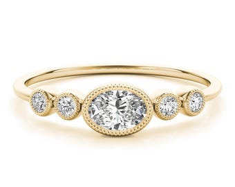 May include: A gold ring featuring an oval-cut diamond center stone, framed by a beaded bezel. Flanking the center are four smaller round diamonds, two on each side. The band is a thin, polished gold.