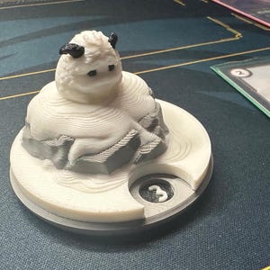 May include: A 3D-printed white and gray game piece featuring a stylized creature with black horns and eyes. The piece sits on a circular base with a number 3. A card game is in the background.
