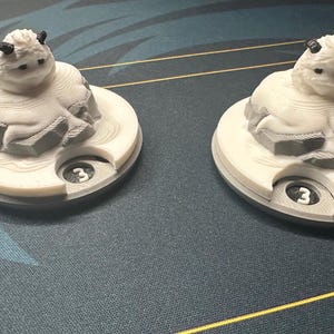 May include: Two white and gray game pieces, each featuring a stylized creature with small horns, resting on a circular base. The base has a cutout with the number "3" visible. The pieces are on a patterned surface.