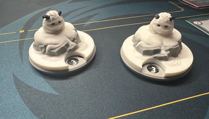 May include: Two white and gray game pieces, each featuring a stylized creature with small horns, resting on a circular base. The base has a cutout with the number "3" visible. The pieces are on a patterned surface.