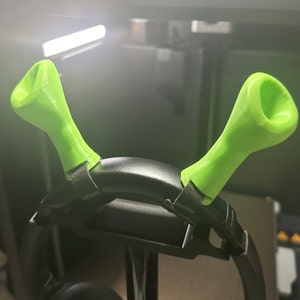 Puede incluir: Un par de ganchos para auriculares impresos en 3D de color verde, unidos a una diadema negra. Los ganchos están diseñados para sujetar los auriculares en su lugar.