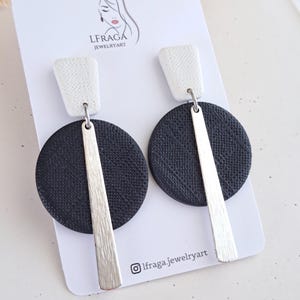 Può includere: Un paio di orecchini moderni con una parte superiore rettangolare bianca e una parte inferiore circolare nera. Una barra d'argento collega le due parti. Gli orecchini sono esposti su una carta bianca con il testo "LFRAGA JEWELRYART" e un logo.