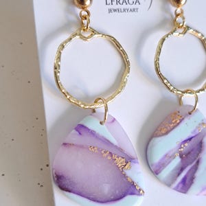 May include: A pair of gold-toned earrings with a textured hoop design and a small gold ball at the top. The earrings feature a marbled, triangular pendant in shades of purple, mint green, and gold. The brand name "LFRAGA JEWELRYART" is visible.