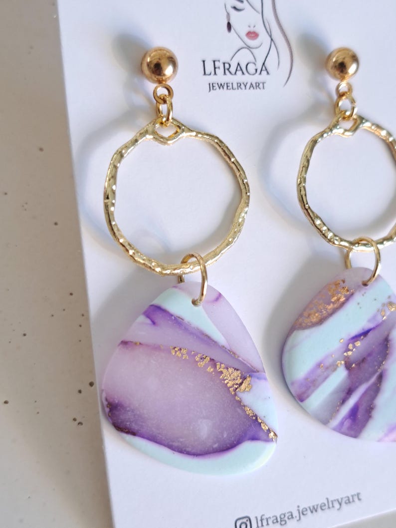 May include: A pair of gold-toned earrings with a textured hoop design and a small gold ball at the top. The earrings feature a marbled, triangular pendant in shades of purple, mint green, and gold. The brand name "LFRAGA JEWELRYART" is visible.