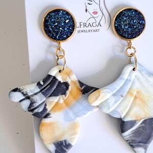 May include: A pair of handmade earrings. Each earring features a gold-colored circular top with a dark blue, glittery center. The lower portion is a marbled design in white, black, and yellow, with a wing-like shape. The earrings are displayed on a white card.