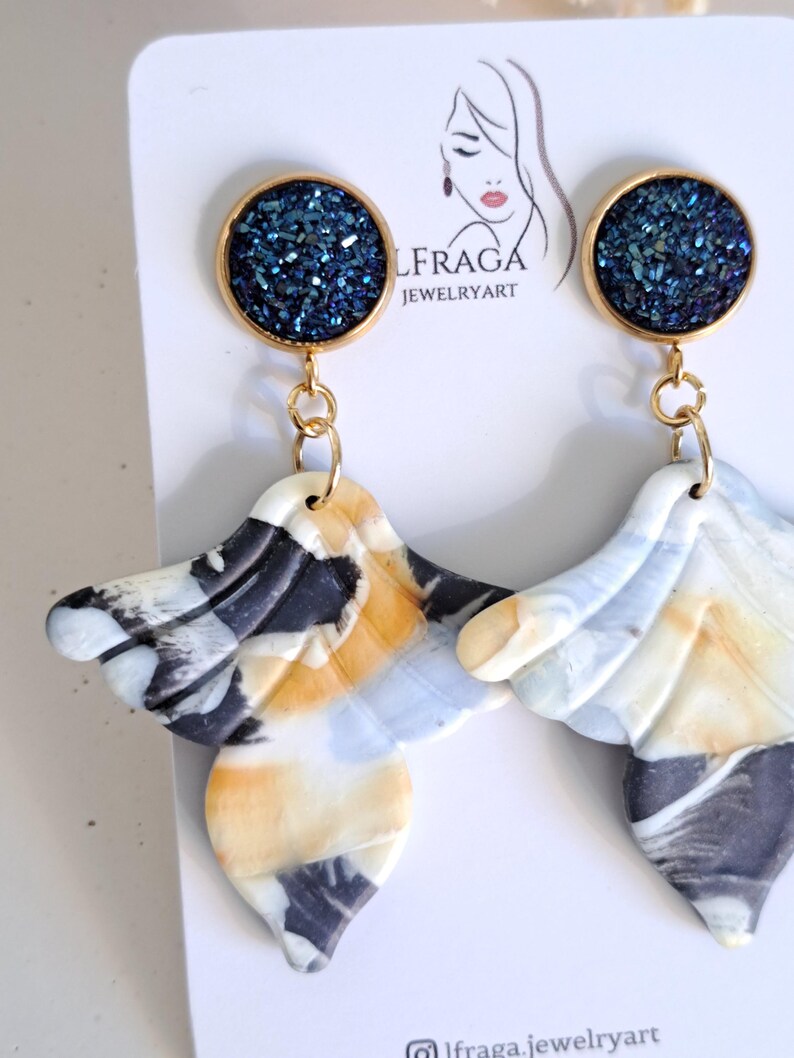 May include: A pair of handmade earrings. Each earring features a gold-colored circular top with a dark blue, glittery center. The lower portion is a marbled design in white, black, and yellow, with a wing-like shape. The earrings are displayed on a white card.