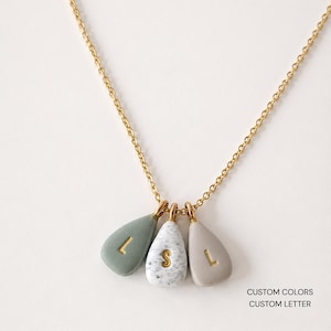 Pode incluir: Um colar de corrente dourada com três pingentes em forma de gota nas cores verde sálvia, branco salpicado e bege. Cada pingente é marcado com uma letra dourada: L, S e L. O texto CUSTOM COLORS CUSTOM LETTER está no canto inferior direito.