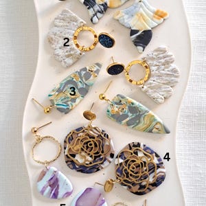 May include: A collection of handmade earrings in various designs. The earrings feature a mix of colors including blue, white, gold, and purple. Some earrings have a marbled effect, while others have a floral design. The earrings are displayed on a white, wavy surface.