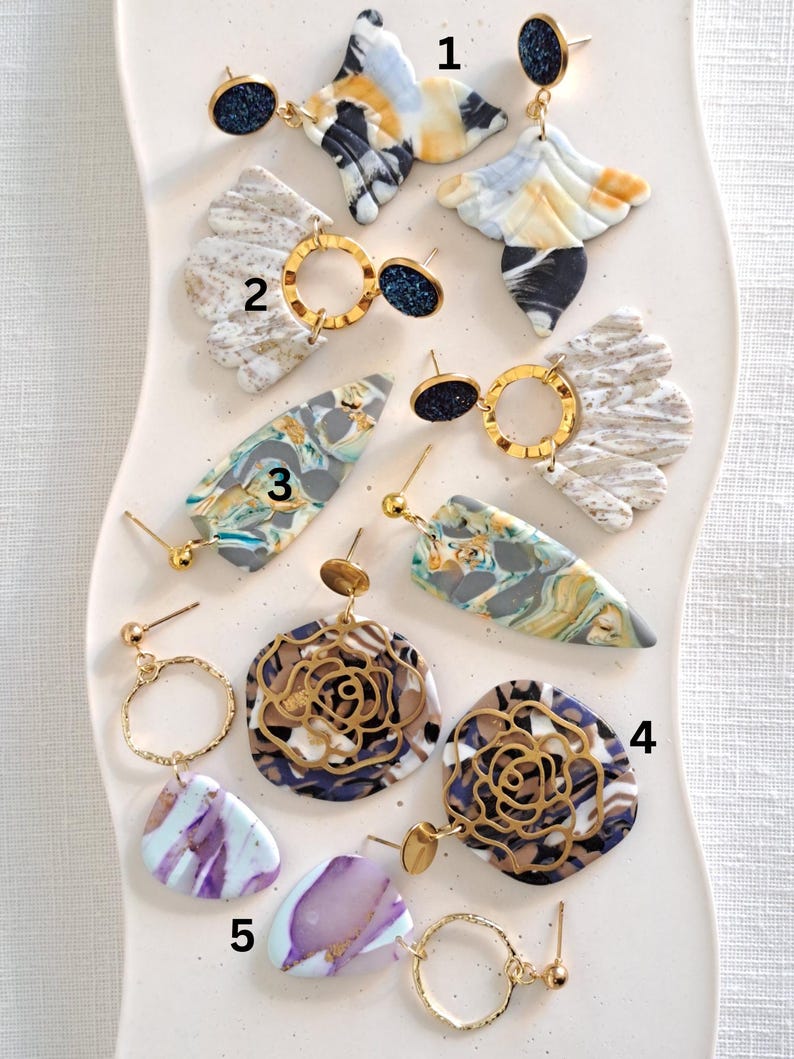 May include: A collection of handmade earrings in various designs. The earrings feature a mix of colors including blue, white, gold, and purple. Some earrings have a marbled effect, while others have a floral design. The earrings are displayed on a white, wavy surface.