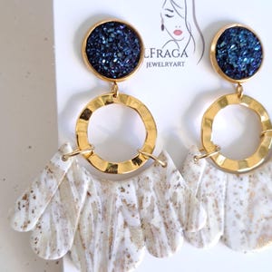 May include: A pair of statement earrings featuring a dark blue, glittery circular top, connected to a gold-colored ring and a white and beige marbled fan-shaped bottom. The earrings are displayed on a white card with the brand name "LFRAGA JEWELRYART".