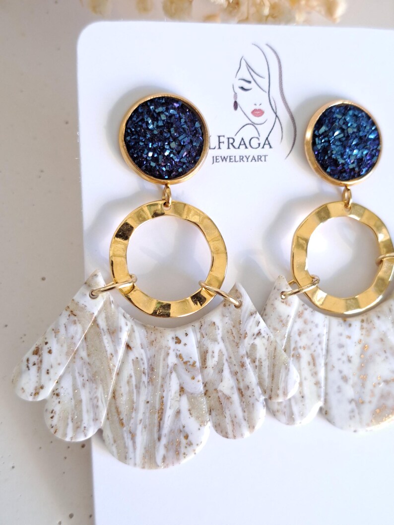 May include: A pair of statement earrings featuring a dark blue, glittery circular top, connected to a gold-colored ring and a white and beige marbled fan-shaped bottom. The earrings are displayed on a white card with the brand name "LFRAGA JEWELRYART".