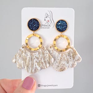 May include: A pair of dangle earrings with a navy blue glitter top, gold-colored ring, and a fan-shaped bottom with a white and beige marbled pattern. The earrings are displayed on a white card with the text "LFRAGA JEWELRYART".