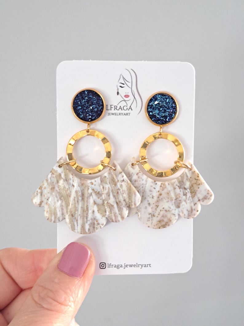 May include: A pair of dangle earrings with a navy blue glitter top, gold-colored ring, and a fan-shaped bottom with a white and beige marbled pattern. The earrings are displayed on a white card with the text "LFRAGA JEWELRYART".
