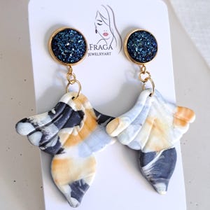 May include: A pair of handmade earrings with a gold-colored top and a marbled design in shades of white, black, and orange. The top of each earring features a dark blue, glittery circle. The earrings are displayed on a white card with the text "IFRAGA JEWELRYART".