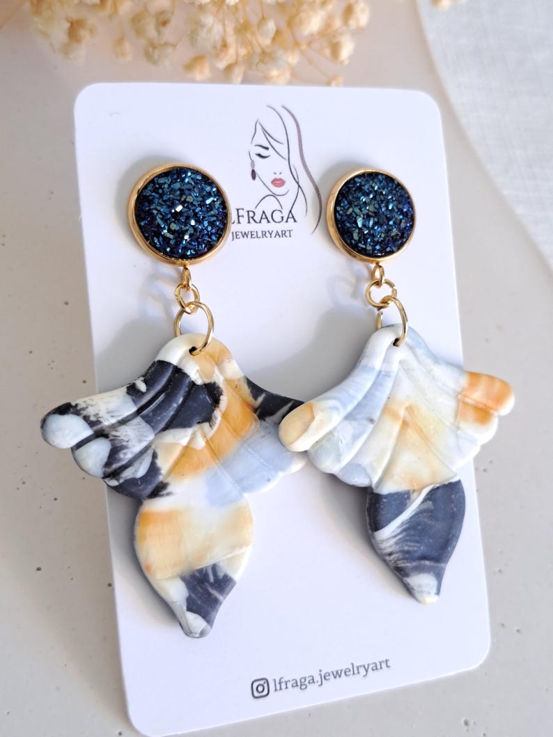 May include: A pair of handmade earrings with a gold-colored top and a marbled design in shades of white, black, and orange. The top of each earring features a dark blue, glittery circle. The earrings are displayed on a white card with the text "IFRAGA JEWELRYART".