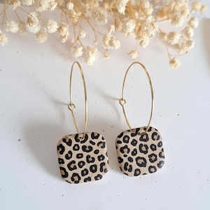 May include: A pair of gold hoop earrings with square, leopard print pendants. The pendants feature a tan background with black spots. The earrings are displayed on a white surface with dried flowers in the background.