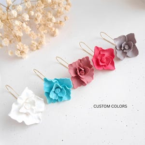 Puede incluir: Un conjunto de cinco pendientes con forma de flor en varios colores: blanco, turquesa, rosa empolvado, rosa brillante y gris. Cada pendiente tiene un gancho de alambre dorado. El texto "CUSTOM COLORS" es visible.