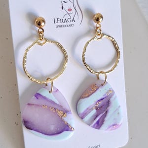 May include: A pair of gold-colored earrings with a circular design and a teardrop-shaped pendant. The pendants feature a marbled pattern in shades of purple, light blue, and gold. The earrings are displayed on a white card with the text "LFRAGA JEWELRYART".
