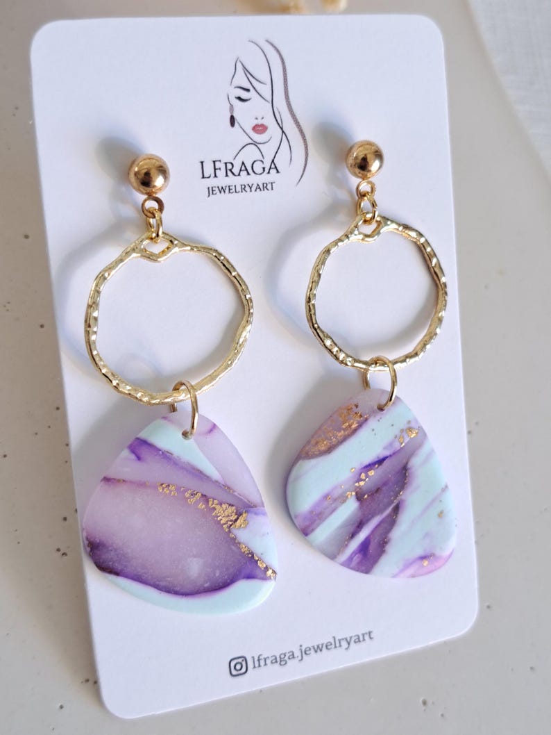 May include: A pair of gold-colored earrings with a circular design and a teardrop-shaped pendant. The pendants feature a marbled pattern in shades of purple, light blue, and gold. The earrings are displayed on a white card with the text "LFRAGA JEWELRYART".