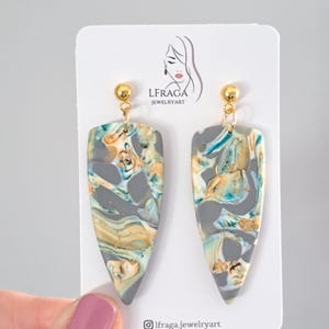 May include: A pair of handmade earrings with a marbled design. The earrings feature a gray base with swirls of teal, gold, and orange. The earrings are teardrop-shaped and have gold-colored ball studs. The earrings are displayed on a white card with the text "LFRAGA JEWELRYART".