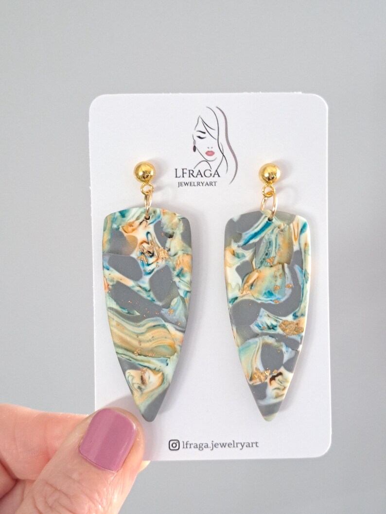 May include: A pair of handmade earrings with a marbled design. The earrings feature a gray base with swirls of teal, gold, and orange. The earrings are teardrop-shaped and have gold-colored ball studs. The earrings are displayed on a white card with the text "LFRAGA JEWELRYART".