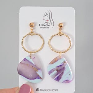 May include: A pair of gold-toned hoop earrings with abstract, marbled pendants in shades of purple, blue, and gold. The earrings are displayed on a white card with the brand logo.