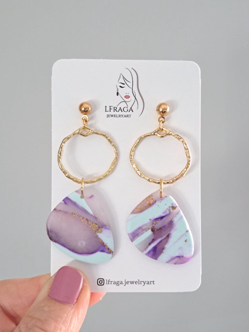 May include: A pair of gold-toned hoop earrings with abstract, marbled pendants in shades of purple, blue, and gold. The earrings are displayed on a white card with the brand logo.