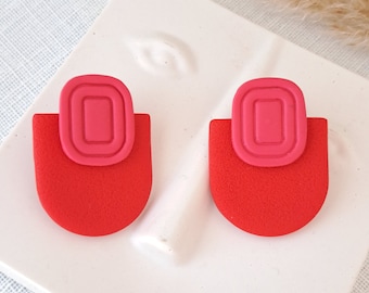 Puede incluir: Un par de pendientes de botón con una base semicircular de color rojo brillante rematada con un elemento rectangular rosa más pequeño y elevado que presenta un diseño de ranuras concéntricas. Tienen un acabado mate y texturizado.