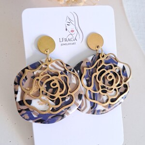 May include: A pair of statement earrings featuring a gold-colored rose design overlaying a marbled pattern in shades of blue, brown, and white. The earrings are attached to a gold-colored circular stud. The earrings are displayed on a white card with the text "LFRAGA JEWELRYART".