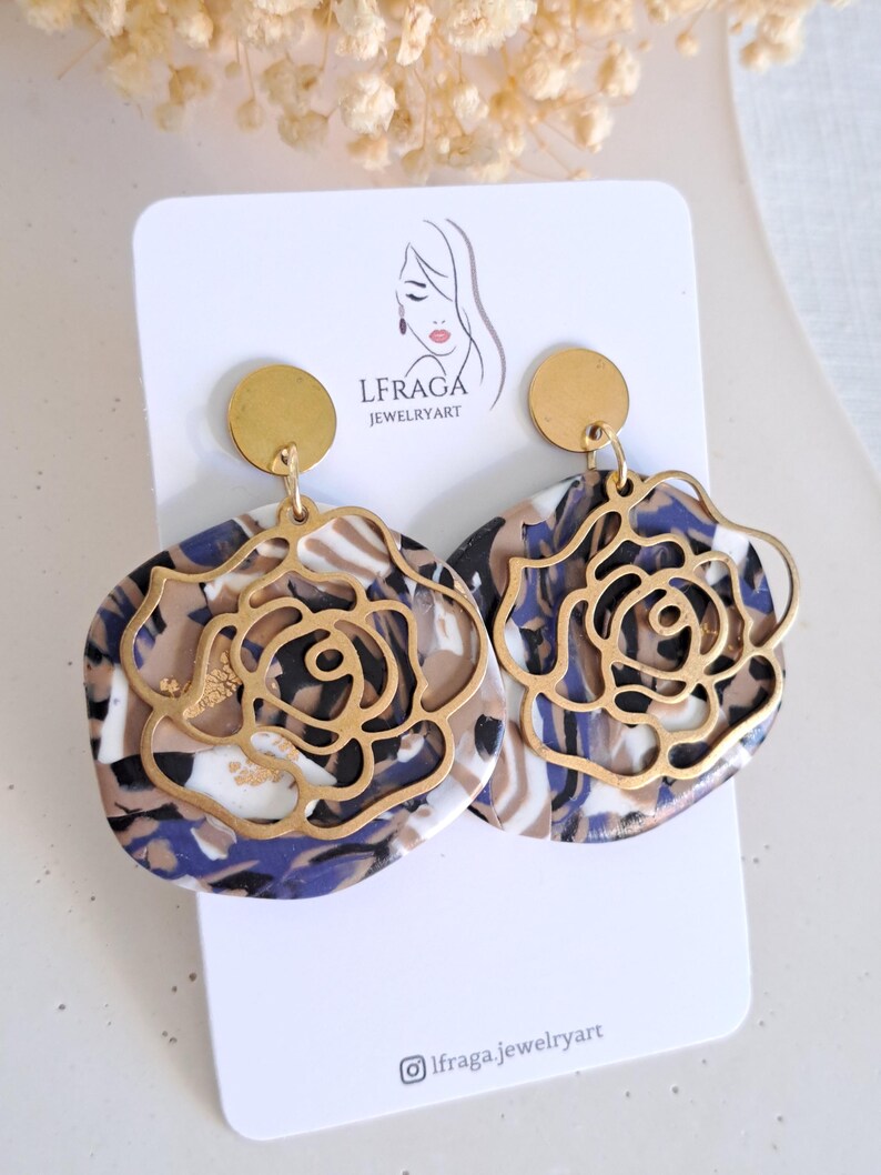May include: A pair of statement earrings featuring a gold-colored rose design overlaying a marbled pattern in shades of blue, brown, and white. The earrings are attached to a gold-colored circular stud. The earrings are displayed on a white card with the text "LFRAGA JEWELRYART".