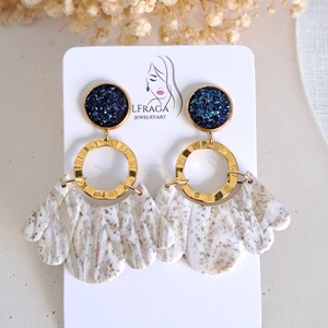 May include: A pair of dangle earrings with a gold-tone metal design. The earrings feature a dark blue, glittery top, a gold hoop, and a white and brown marbled fan-shaped bottom. The earrings are displayed on a white card with a logo.