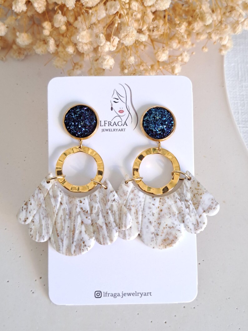 May include: A pair of dangle earrings with a gold-tone metal design. The earrings feature a dark blue, glittery top, a gold hoop, and a white and brown marbled fan-shaped bottom. The earrings are displayed on a white card with a logo.