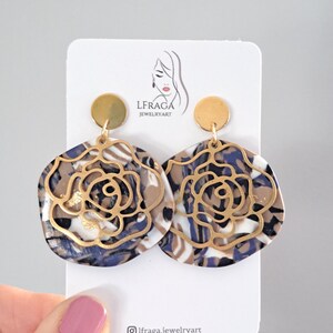 May include: A pair of statement earrings featuring a floral design. The earrings have a gold-colored metal overlay on a marbled base of white, navy, and brown. The earrings are displayed on a white card with the text "LFRAGA JEWELRYART".